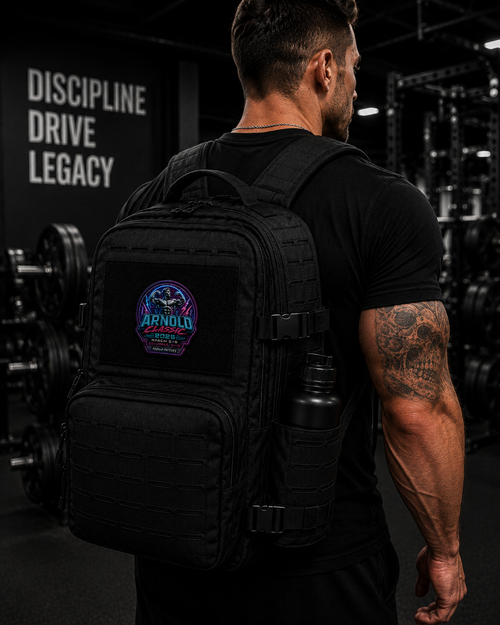 A man in a black t-shirt seen from behind in a gym, wearing a black tactical backpack with a circular 'The Arnold' logo patch on the front pocket and a water bottle in the side pocket.